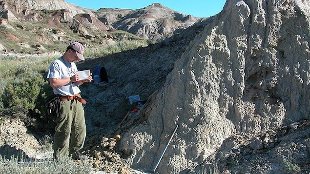 Un homme debout en train de mener des fouilles dans un paysage de montagne prend des notes dans un carnet. 