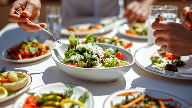 Une table est couverte de plusieurs plats de salades diverses et une femme se sert dans l'une d'elles. 