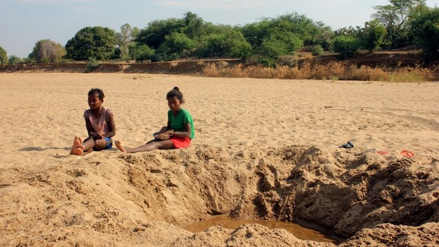 Deux fillettes sont assises par terre à côté d'un trou creusé dans un champ de sable au fond duquel apparaît un peu d'eau. 
