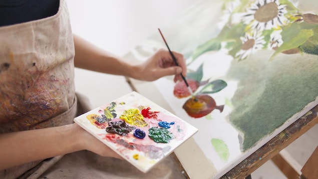 Gros plan sur une femme, pinceau à la main, penchée sur une toile qui représente des fleurs et des fruits.