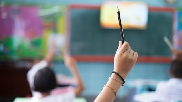 Un enfant dans une salle de classe lève le bras en tenant un crayon dans sa main.