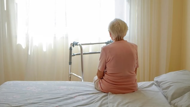 Une femme âgée est assise sur un lit dans une chambre.