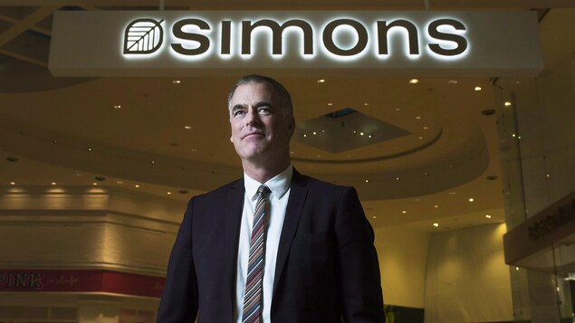 Peter Simons pose en costume devant l'enseigne lumineuse d'un magasin Simons.
