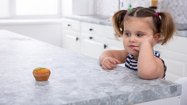 À gauche, un petit gâteau sur un comptoir et, à droite, une jeune fille avec des couettes qui le regarde.