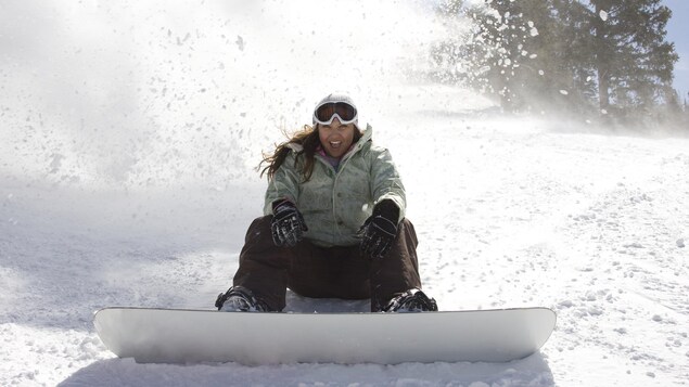 Une planchiste sourit, la bouche ouverte, assise dans une pente après un freinage qui soulève la neige autour d'elle. 
