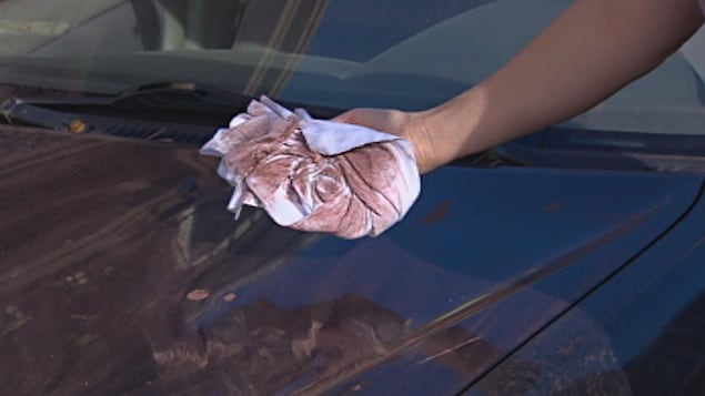 Une main brandit un chiffon blanc maculé de poussière rouge après avoir été utilisé pour essuyer le capot d’une voiture.