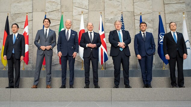 Les dirigeants du G7 prennent la pose pour une photo de famille : sept hommes en complet se tiennent debout alignés en une même rangée devant des drapeaux. 
