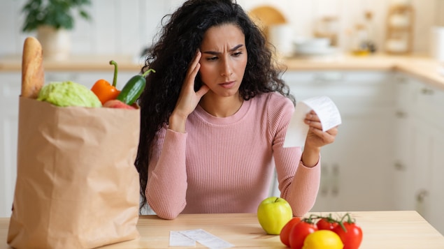 Una mujer sentada en su cocina mira con detalle su factura d'épicerie après avoir fait des courses. Un sac en papier brun avec des aliments est posé sur la table.