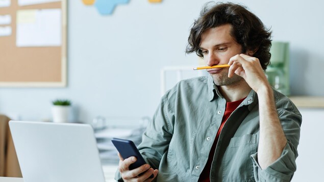 Portrait d'une personne qui s'amuse avec un stylo tout en regardant sont téléphone plutôt que de travailler sur son ordinateur.