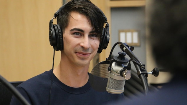 Un homme, casque d'écoute sur la tête, regarde en souriant dans la direction de l'animateur dans un studio de radio. 