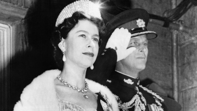 Dans cette photo en noir et blanc, la reine se tient debout devant son mari qui fait un salut de la main.