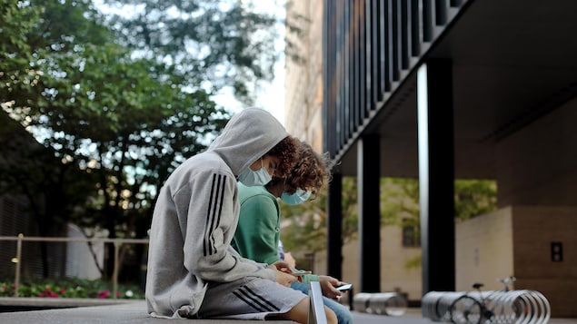 Deux ados masqués regardent leur téléphone, assis sur du mobilier urbain à l'extérieur d'une école.  
