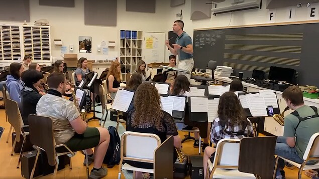 Richard Piette est debout devant une salle de classe pleines d'élèves avec des instruments, surtout des cuivres. Il a dans ses mains une baguette de conducteur.