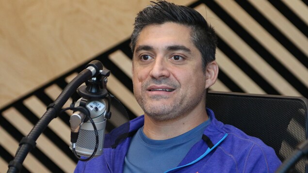 Un homme esquisse un sourire en coin derrière un micro, dans un studio de radio. 