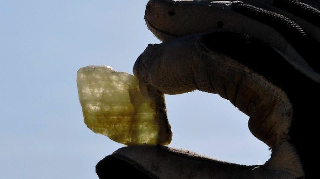 On voit une main gantée tenant un cristal de spodumène devant un ciel ensoleillé.