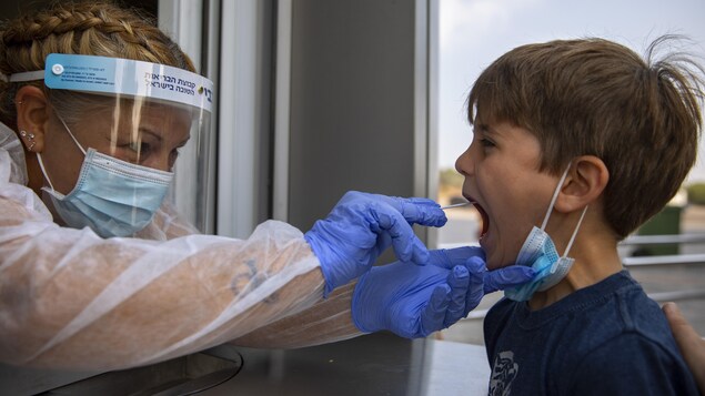 Une infirmière avec une visière et des gants administre un test covid à un jeune garçon en introduisant un écouvillon dans sa bouche.