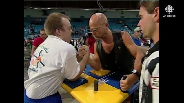 Deux athlètes s'affrontent lors d'une compétition de tir au poignet sous les yeux attentifs d'un arbitre. 