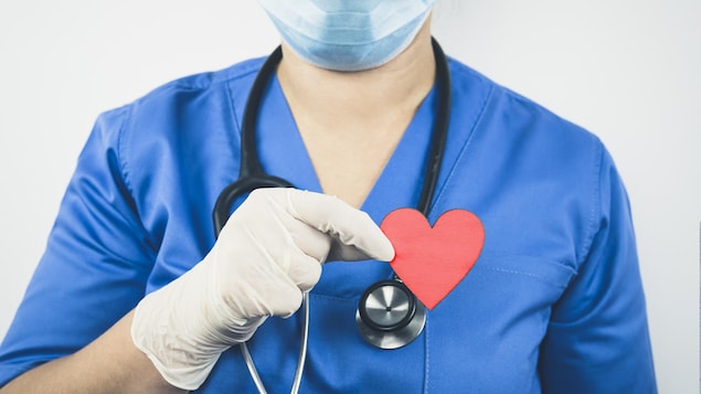 Une médecin portant un masque chirurgical et des gants en plastique tient un cœur entre ses doigts.