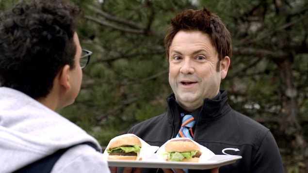 Le comédien sourit en tenant un plateau avec deux sandwiches.