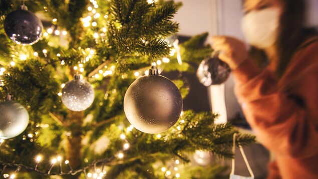 Une femme portant un masque décore un sapin de Noël.