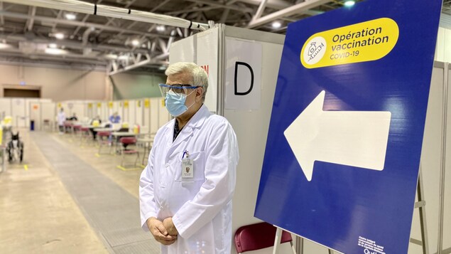 Un homme en blouse et masque se tient debout dans un hangar devant une pancarte "opération vaccination COVID-19".