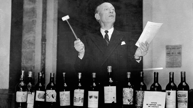 Un homme avec un marteau et une feuille devant une table avec une douzaine de bouteilles de vin vendues aux enchères.