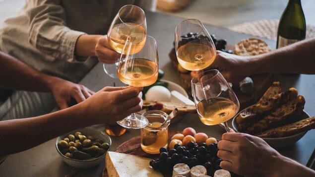 Quatre personne trinque avec des verres de vin au-dessus d'une table avec un plateau composé de fromages, de pains et de fruits et de divers condiments. 