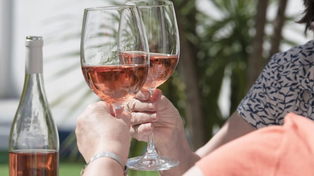Les mains de deux personnes qui se font un toast avec un verre de vin rosé. Elles sont dehors, l'été, et il y a une bouteille sur la table.