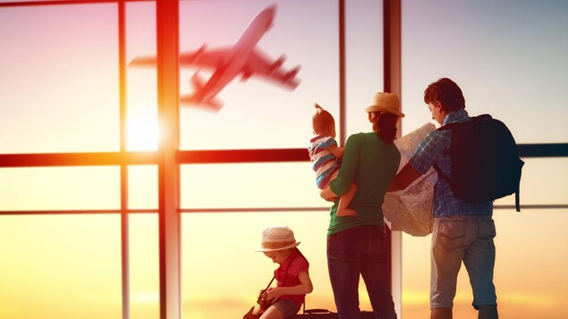 Une famille, une jeune fille assise sur une valise, une mère qui porte un jeune enfant dans ses bras, et un père qui consulte une carte routière se trouve devant les vitres d’un aéroport alors qu’un avion décolle dehors.