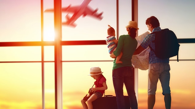 Une famille, une jeune fille assise sur une valise, une mère qui porte un jeune enfant dans ses bras, et un père qui consulte une carte routière se trouve devant les vitres d’un aéroport alors qu’un avion décolle dehors.