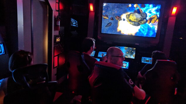 Vue de dos des participants au jeu d'évasion Proxima Command assis dans un simulateur de salle de contrôle.