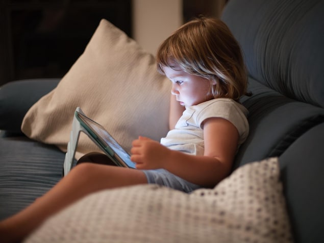 Une jeune enfant assise sur un canapé en train de regarder l'écran d'une tablette électronique.