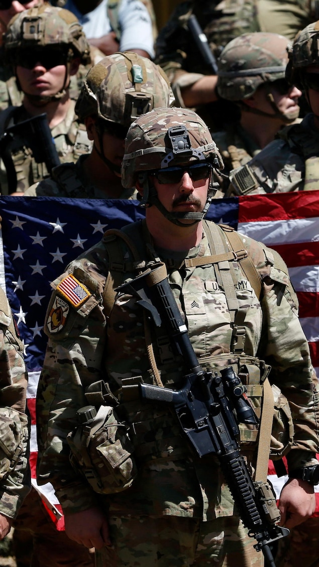 Des soldats américains armés devant un drapeau des États-Unis tenu par d'autres soldats.