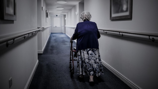 Une vieille personne de dos marche dans un couloir avec une marchette.