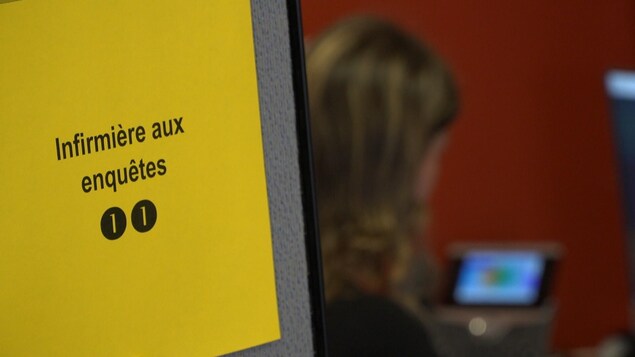 Gros plan sur une feuille de papier jaune sur laquelle sont écrits les mots « infirmière aux enquêtes ». La feuille est affichée sur une cloison délimitant un espace de travail. On aperçoit, à l’arrière-plan, une femme parlant au téléphone.