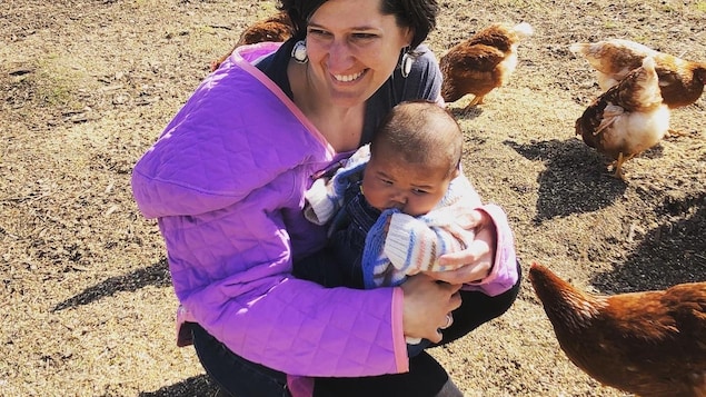 Jessica tient la petite Joy dans ses bras alors qu'elles sont entourées de poules dans un enclos.