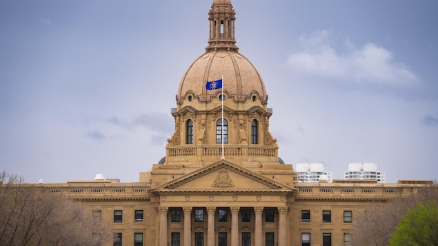 Gros bâtiment beige avec un drapeau de l'Alberta bleu qui flotte au vent. Ciel gris foncé avec nuages plus pâles. Peu de feuille aux arbres. 15 mai 2024.