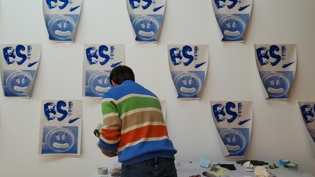 Un artiste devant un mur où sont collées plusieurs affiches sur lesquelles on peut lire BSL.