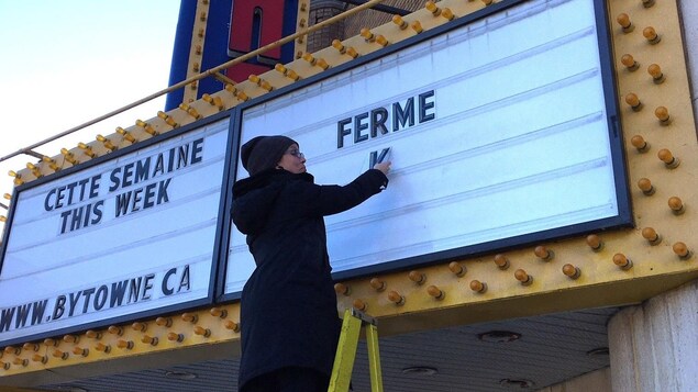 Une femme en tain d'installer les lettres du mot « ferme » sur l'affiche d'un cinéma.