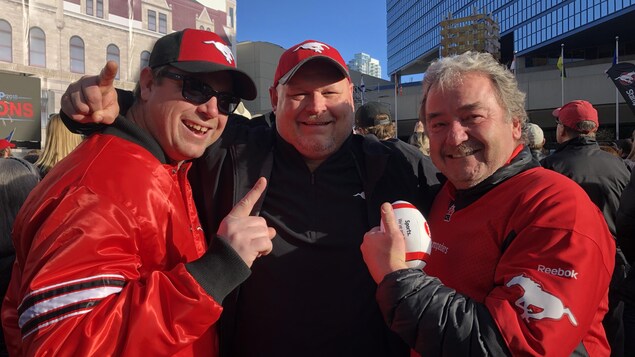Trois hommes habillés aux couleurs des Stampeders sourient à la caméra. Trois hommes habillés aux couleurs des Stampeders sourient à la caméra.