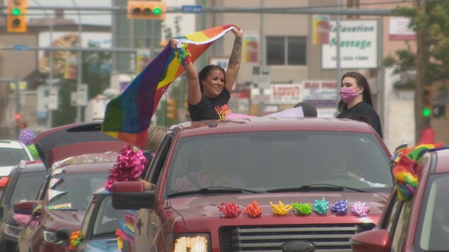 Une femme tenant un drapeau LGBTQ+ debout dans un pickup décoré. Un autre femme à côté avec un masque sur le visage. File de voitures décorées aux couleurs de l'arc en ciel.