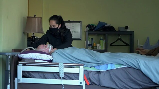 Cristina David s'occupe de son mari, Simon, allongé sur un lit médical et portant un masque d'assistance respiratoire.
