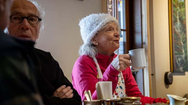 Deux aînés à la table d'un restaurant. À gauche un homme parle avec son voisin (qui n'est pas sur la photo). À droite, une femme sourit, une tasse de café à la main.