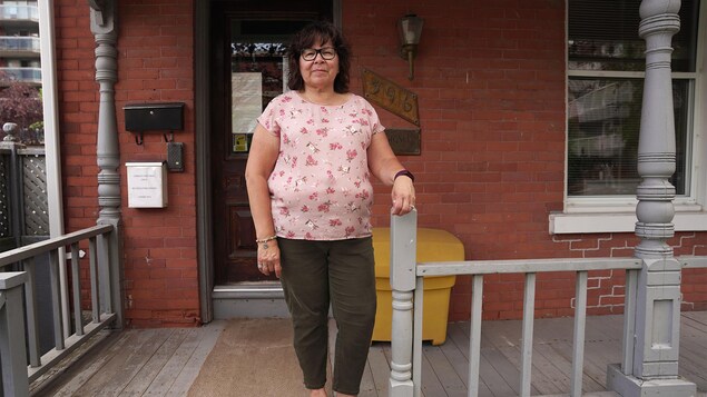 Une femme souriante debout devant une porte de maison. Son bras gauche est apposé sur une rampe.