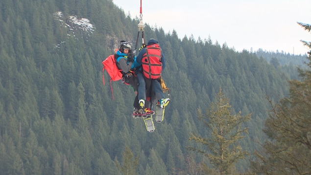 Deux sauveteurs portent secours à une personne blessée à ski. Les trois sont hélitreuillés et suspendus au-dessus du vide.