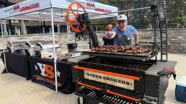 Graham Sherman montre une bière derrière un barbecue rempli de morceaux de poulet. 