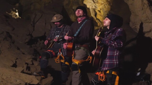 Trois musiciens vêtus d'équpiement de spéléologie jouent de la guitare dans une grotte. 