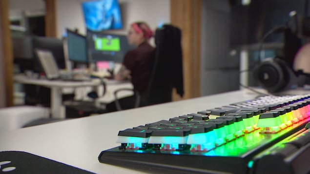 Un clavier s'illumine de couleurs alors qu'une femme travaille à un ordinateur en arrière-plan. 