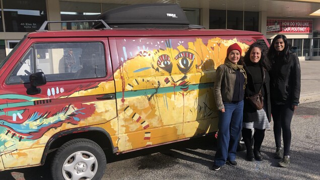 Trois femmes posent devant une fourgonnette Westfalia 1985, le visage d'une femme est peinte de façon abstraite sur le véhicule.
