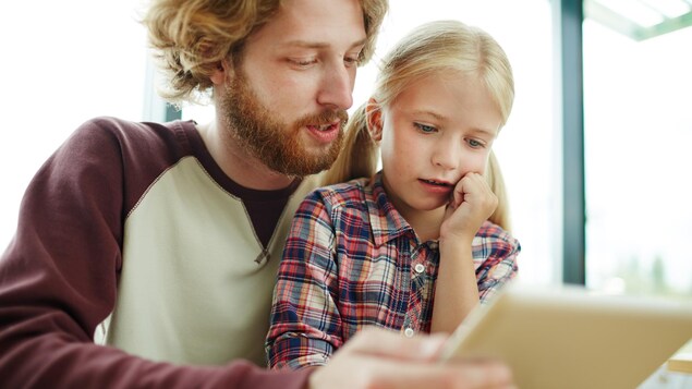 Un père tient une tablette électronique et la montre à sa fille. 